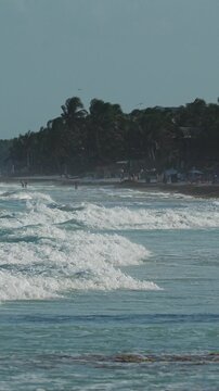 Tulum, Yucatan, Mexico - 15 March 2025: Crashing waves roll toward shore. Clear sky above distant palm trees. Blue tent sits near the water. People relax and enjoy the view. Gentle turquoise water gli