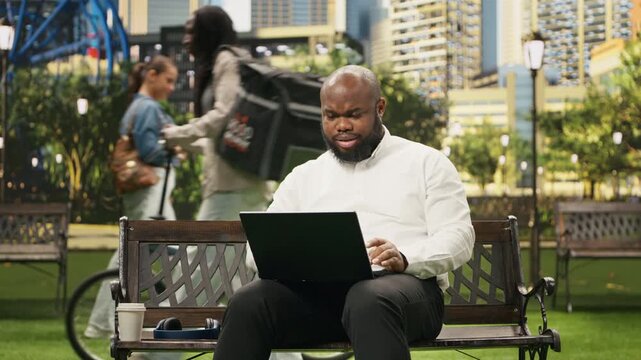 Excited businessman raises clenched fists while looking at a laptop on a park bench, celebrating a positive result and success during flexible outdoor work.