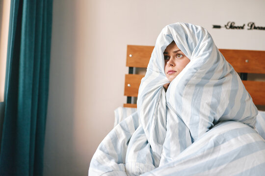 Sad woman wrapped in a blanket sitting on a bed in a bedroom. Concept of comfort, illness, and cold. Soft lighting creates a cozy yet lonely mood.