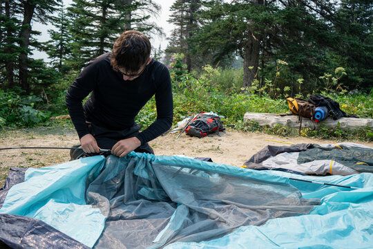 Young man setting up a blue tent in a pine forest during a camping trip in the mountains