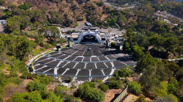 Cinematic aerial view of famous open air amphitheater in Hollywood Hills. Empty seating rows and white stage shell surrounded by green trees. Sunny summer day in Los Angeles California. 