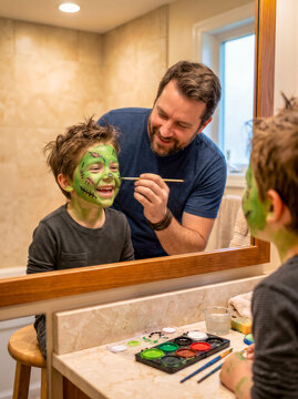 Cheerful father applying green monster makeup face paint on smiling young son in bright bathroom mirror for Halloween celebration. American carnival for Halloween celebrating concept with copy space.