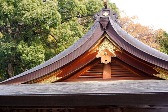 日本の伝統的な建築物の優美な屋根のディテール。金色の装飾金具と猪目の彫刻が施された木製の破風、銅板葺きの屋根が秋の木々を背景に引き立つ。