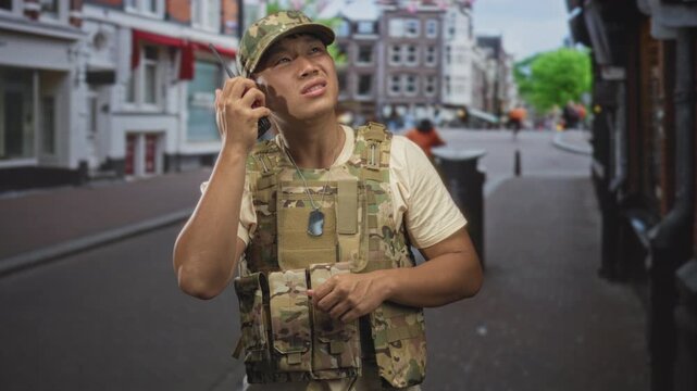 Man soldier in camouflage vest holds radio to ear on a street, dog tags visible, listening and squinting; alert duty.