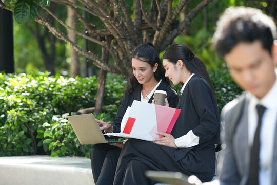 A group of three smartly dressed coworkers briefing the team from a tablet beside a bench at a public bench in the business district, a modern picture of teamwork in a green city district