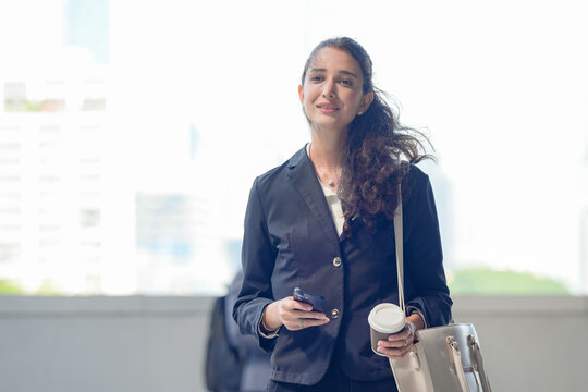 A woman in leadership wearing business attire checking messages on a smartphone with coffee in hand on a skywalk above the business district, showing how work happens beyond the meeting room