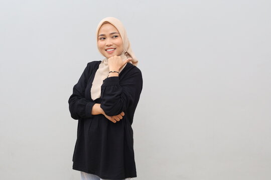 A beautiful young Asian woman wearing a hijab smiles while pointing her thumb to the empty space beside her and glancing sideways, indicating an advertisement, standing against a white background.