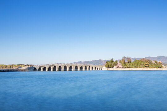 A multi-arch stone bridge spans a calm lake, backed by mountains and trees under a clear blue sky.