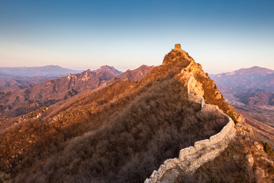 Ancient stone Great Wall winds along a mountain peak at sunrise, with distant mountain ranges under clear sky.