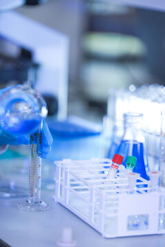 The gloved hands of a medical research scientist pouring from flask into a graduated cylinder inside a busy medical diagnostics laboratory, in a clean and modern laboratory.