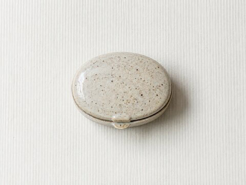 Oval speckled ceramic pillbox on a pale ribbed paper surface with bright overhead daylight