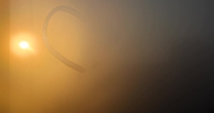 Bright sunlight through a misted glass window and a heart pattern, the orange sun shines behind small droplets of condensation on the glass surface at sunset