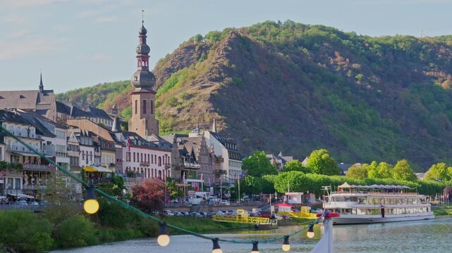 Cinematic video of the historic town of Cochem along the Moselle river featuring its iconic church tower, traditional waterfront architecture, and a string of warm glowing lights hanging in the foregr