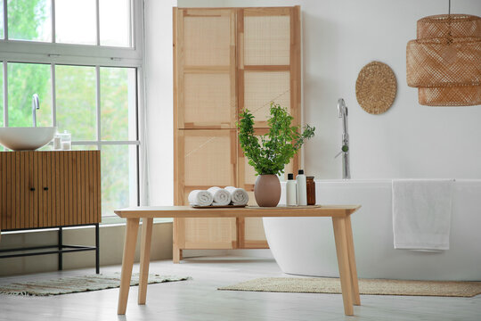 Interior of stylish bathroom with bathtub, folding screen, commode and green blossoming branches in vase on bench near window