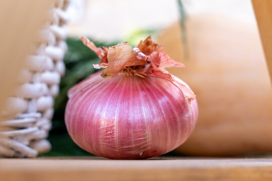 A single fresh red onion with dry papery skin sits on a wooden surface beside a woven basket, a simple closeup of raw produce for cooking.