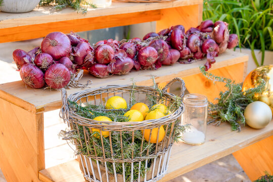 A braided string of red onion bulbs rests on a wooden shelf above a wicker basket filled with lemon fruit and rosemary sprigs, a rustic farmers market display with festive ornaments nearby.