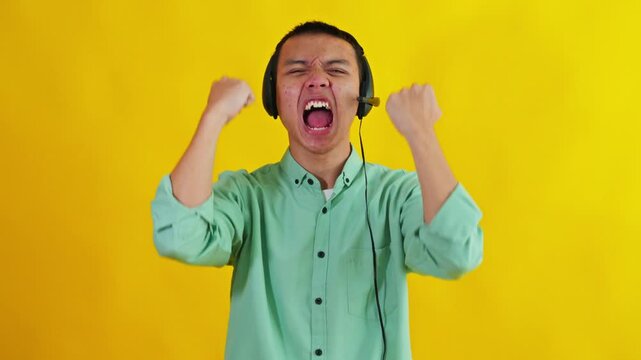 Enthusiastic Asian customer service agent wearing headset celebrating victory on yellow background