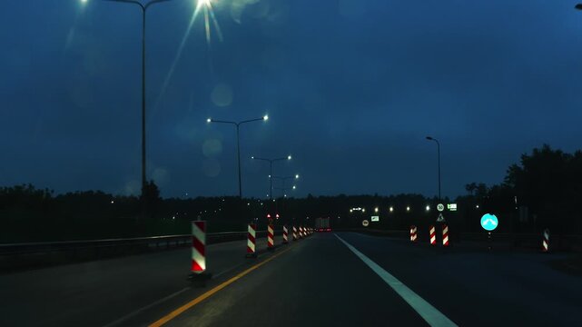 Nighttime highway view with streetlights illuminating traffic signs and construction barriers on a wet road, dark clouds and trees visible in the background