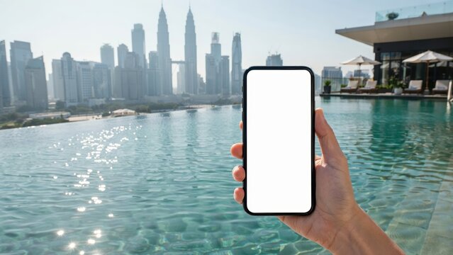 A hand holding a smartphone with a blank screen overlooking a Kuala Lumpur skyline and infinity pool. Ideal for travel apps or mobile digital marketing.