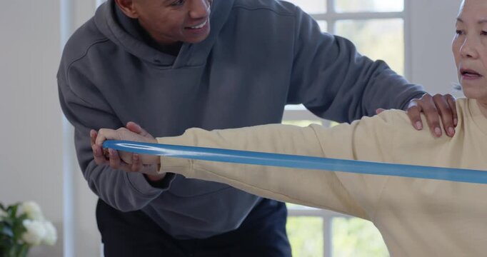 African American man leaning guiding Senior Asian woman extending elastic band for rehab at home