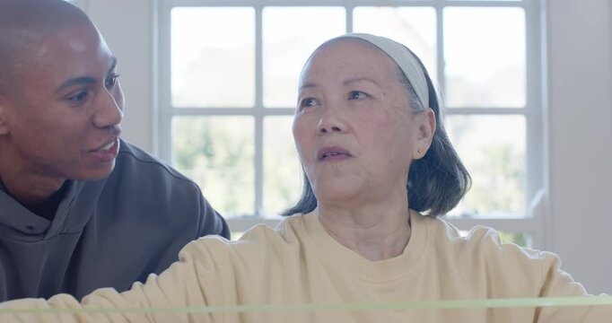 Senior Asian woman holding bands by window, listening as African American man leaning in cues rehab