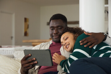 Obraz premium African American couple sitting close on light-colored sofa at home, watching tablet screen