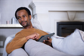 Obraz premium African American man reclining on gray sofa wearing brown quilted top in living room
