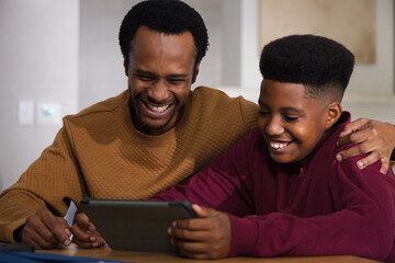 African father, teen son sharing tablet in black case at wooden table at home, maroon zip-up © WavebreakMediaMicro