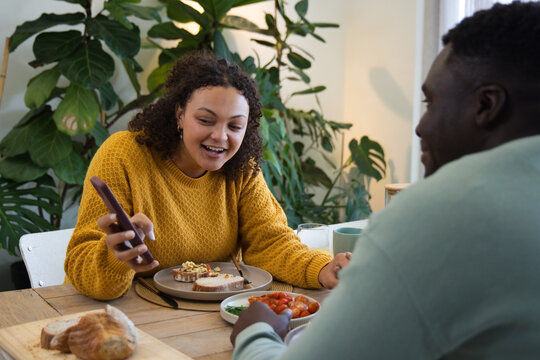 African American couple sitting in dining nook, woman wearing mustard sweater holding smartphone