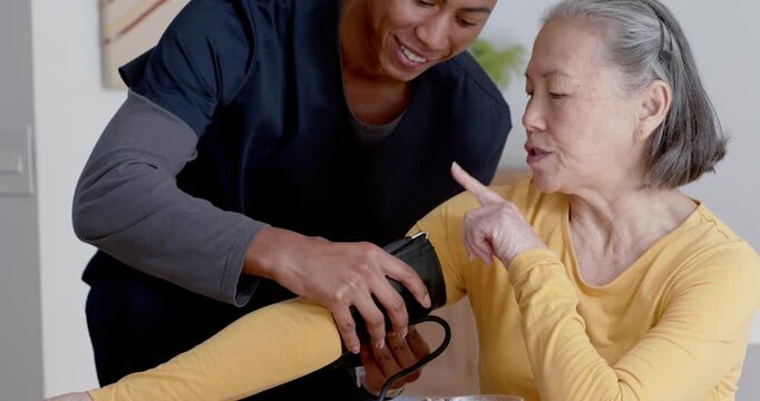 Nurse in scrubs fitting cuff for BP check, Senior Chinese woman extending arm at dining table