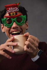 Adult African American man wearing glitter Merry Christmas glasses, holding beige mug, smiling © WavebreakMediaMicro