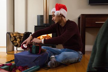 Obraz premium African American man wrapping gifts with ribbons on hardwood floor by wood stove, wearing Santa hat