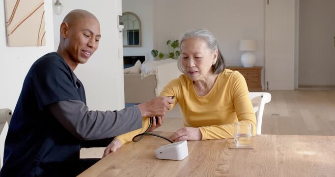 Senior Asian woman and caregiver in scrubs applying cuff at table with monitor measuring BP