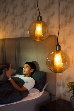 Adult African American male reclining in bed with duvet, holding phone, under amber pendant lamps