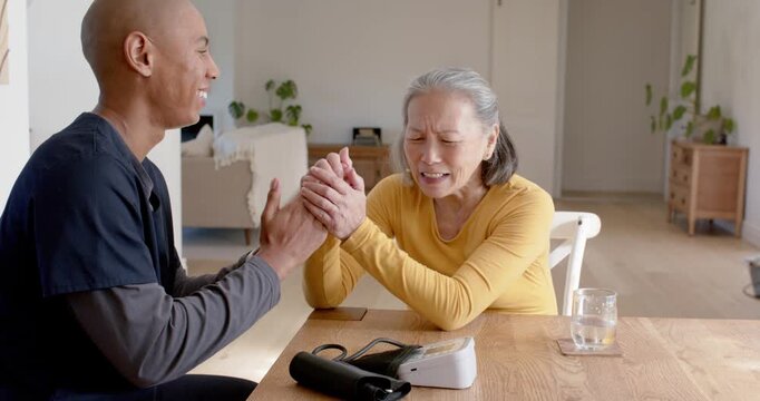 Diverse carer in scrubs guiding senior Asian woman initiating claps for rehab at table with cuff