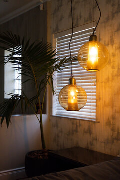 Two amber glass pendant lamps are glowing near window with white blinds, palm casting shadows