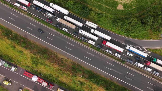 Aerial perspective shows significant traffic jam on a multi-lane highway with cars and trucks, bordered by lush greenery and roadside barriers