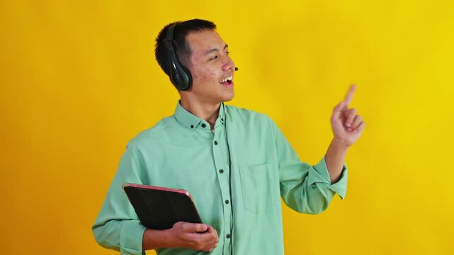 Asian call center agent holding digital tablet and pointing up to side against bright yellow background