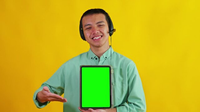 Asian call center agent showing tablet with green screen while speaking on headset on yellow background