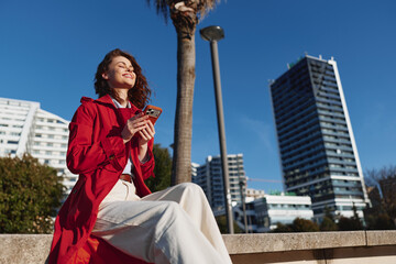 Young woman in red coat sits outdoors on concrete bench holds smartphone enjoys sunny day with...