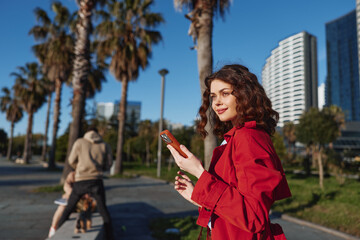 Young woman in red coat holding smartphone outdoors, smiling, sunny day, urban park, palm trees,...