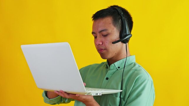 Focused Asian headset operator holding laptop, thinking, and typing data on bright yellow studio background