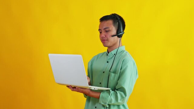 Asian call center agent looking up thoughtfully while holding laptop computer isolated on yellow backdrop