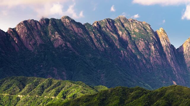 Massive granite mountain ridge covered in green vegetation stands against a blue sky with soft clouds in bright daylight.