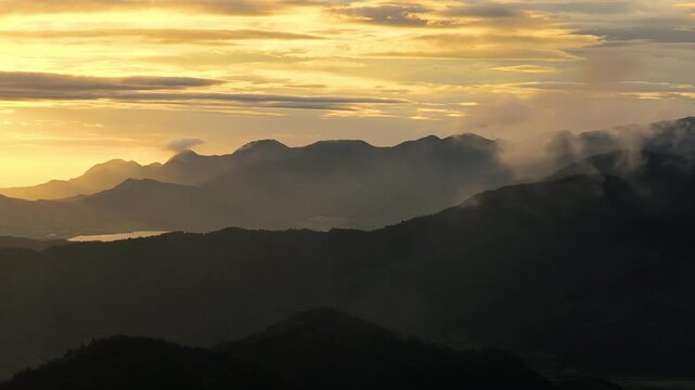 Layered mountain ranges stand illuminated by golden sunset light with soft mist drifting over dark forested ridges in a wide landscape view.