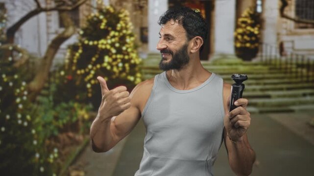 Middle aged hispanic man with beard holding electric shaver and giving thumbs up in front of building steps decorated with string lights; confidence grooming self care.