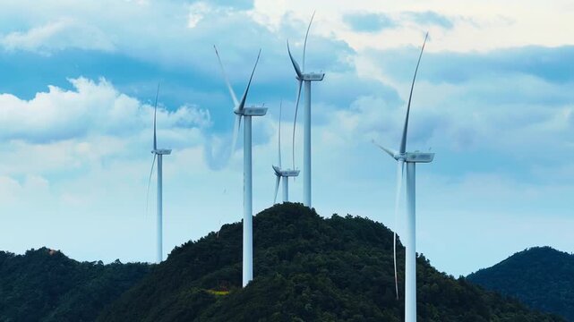 Tall white wind turbines stand on a lush green mountain ridge under a blue sky filled with white clouds in bright daylight.