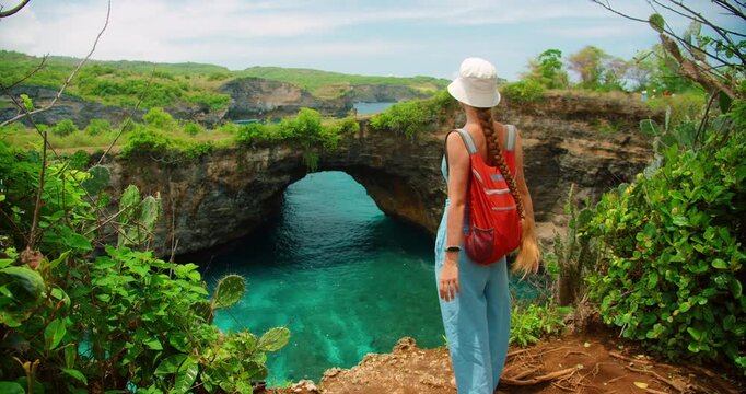 Woman traveler with backpack views ocean through natural archway on tropical cliffside in Broken Beach Nusa Penida Bali