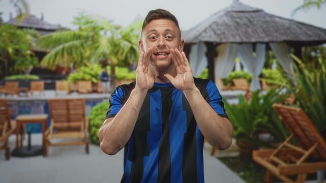 Man cupping hands to mouth while shouting in a resort poolside scene, wearing a blue striped soccer jersey and smiling broadly; excitement support.