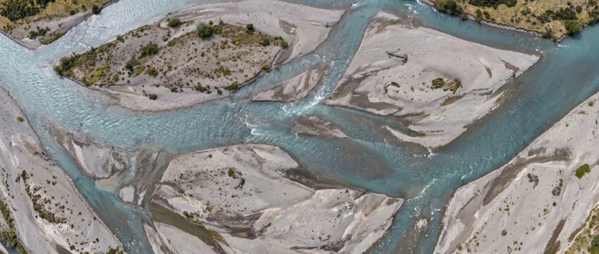 Aerial view of a braided river with turquoise glacial water flowing through light gray gravel riverbeds, sandbars, and sparse green vegetation in a wide valley landscape